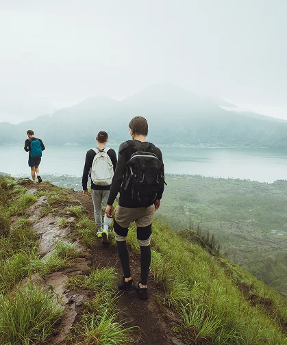 Tři turisté jdou podél zelené, mokré horské stezky s batohy, v pozadí jezero a hory zahalené mlhou.