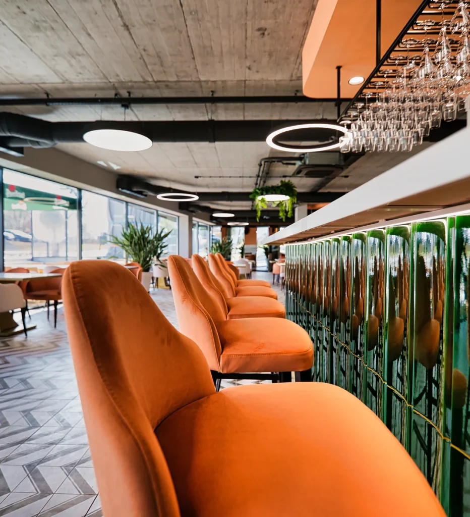 Moderní interiér baru s řadou oranžových barových židlí u zeleného pultu, industriálním stropem a osvětlením z kulatých LED lamp.