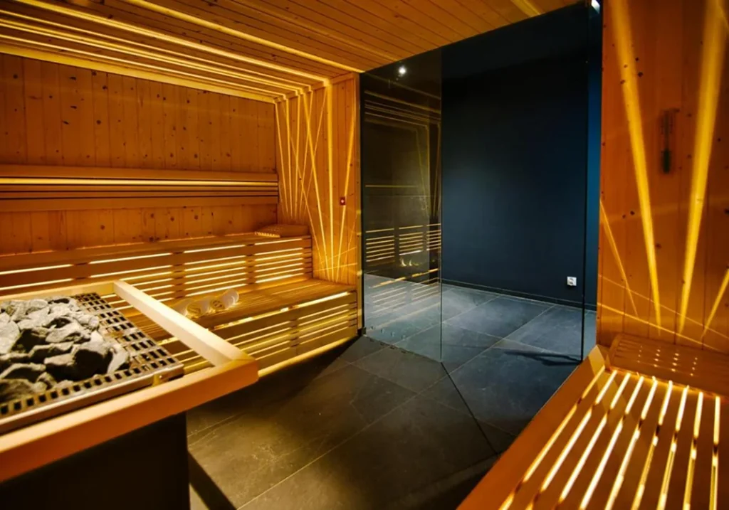 Wnętrze eleganckiej sauny z jasnego drewna z elementami LED, szklaną ścianą i kamiennym piecem, utrzymane w ciepłym, relaksującym świetle.