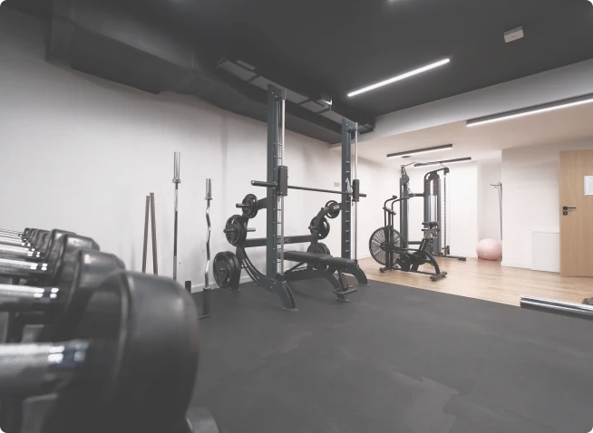 Wnętrze siłowni z czarnymi hantlami, stojakiem do przysiadów, rowerem treningowym i wyciągiem, utrzymane w minimalistycznym stylu z jasnym oświetleniem.
