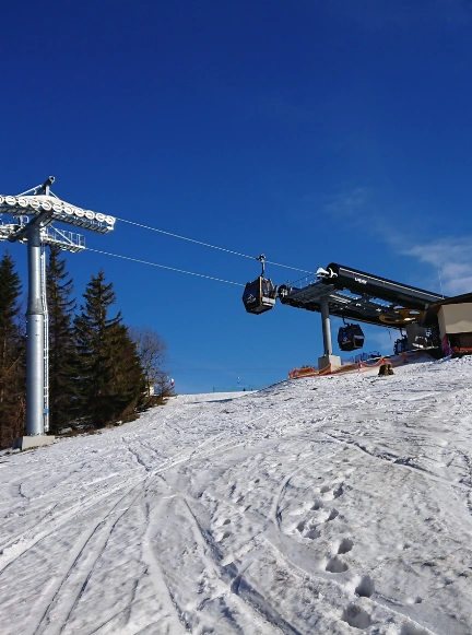Grupa narciarzy zjeżdżająca szeroką, ośnieżoną trasą w słoneczny dzień, w tle widoczna kolejka gondolowa.