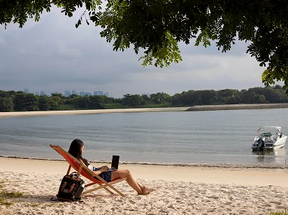 Kobieta siedzi na leżaku z laptopem na piaszczystej plaży, obok plecak, w tle spokojna woda, motorówka i drzewa na brzegu.