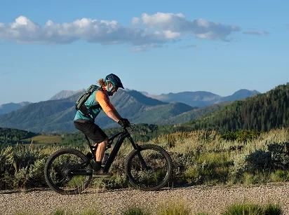 Osoba w kasku i plecaku jedzie na rowerze górskim po żwirowej ścieżce, otoczonej zielenią i górami w tle pod błękitnym niebem.
