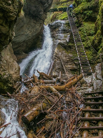Osoba w kasku wspinająca się po długiej, pionowej metalowej drabince tuż obok wodospadu w Słowackim Raju.