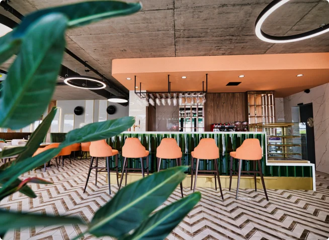 Wnętrze baru z pomarańczowymi hokerami, zieloną ladą i industrialnym sufitem, ozdobione roślinami i geometryczną podłogą w ciepłych tonach.