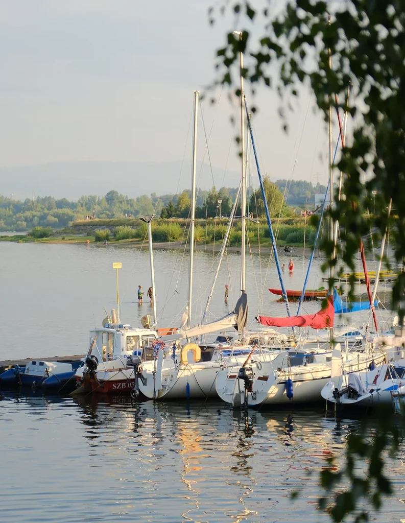 Kilka żaglówek i łodzi motorowych zacumowanych przy pomoście na tle zielonego brzegu jeziora, w ciepłym świetle zachodzącego słońca.
