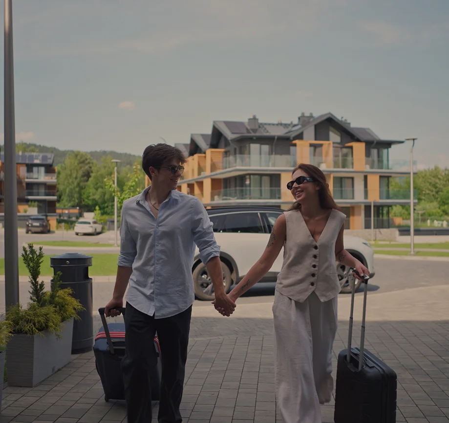 Uśmiechnięta para trzyma się za ręce i wchodzi do nowoczesnego apartamentowca z walizkami na kółkach, w tle widać zaparkowany samochód i zielone otoczenie.